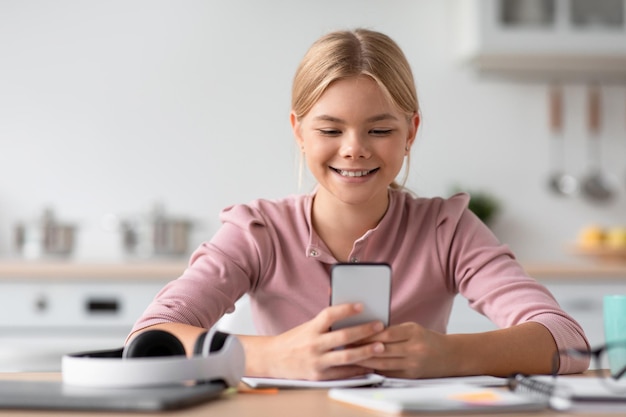 Happy caucasian teenage girl blonde typing on smartphone at table in cozy kitchen interior