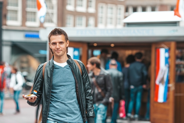 Turista caucasico felice con aringhe fresche con cipolla e bandiera dei paesi bassi ad amsterdam. cibo tradizionale olandese all'aperto