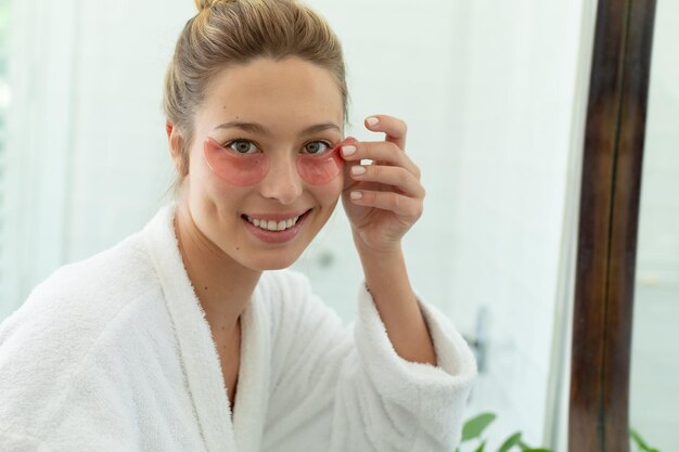 Photo happy caucasian woman in bathrobe putting gel mask under eyes in sunny bathroom