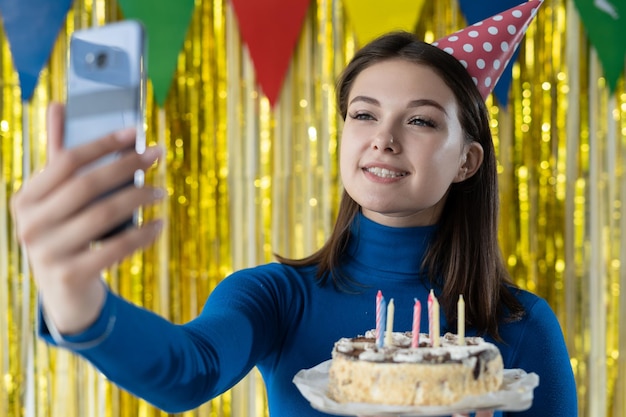 La donna caucasica felice in un vestito blu sta su uno sfondo giallo tiene una torta di compleanno nelle sue mani e fa un selfie sul suo smartphone concetto di festa di compleanno