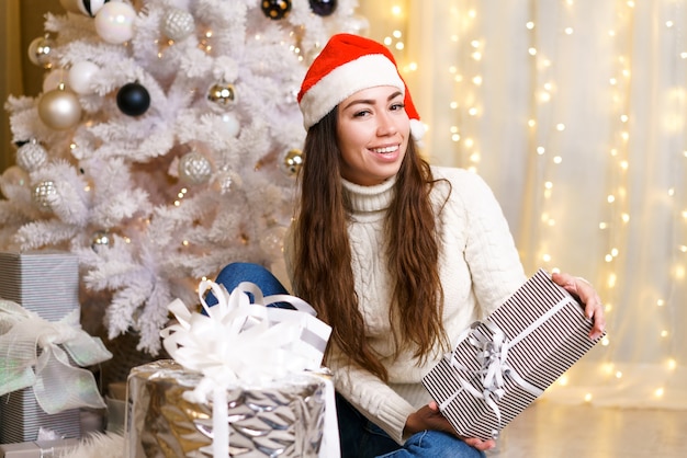 Happy caucasian woman in santa claus hat and light sweater holds a gift box against the backdrop of ...