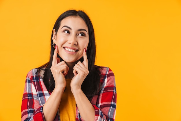 happy caucasian woman smiling and pointing fingers at her cheeks isolated over yellow wall