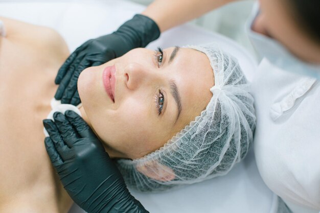 Happy Caucasian woman smiling while unrecognized cosmetologist in rubber gloves touching her neck with cotton pads