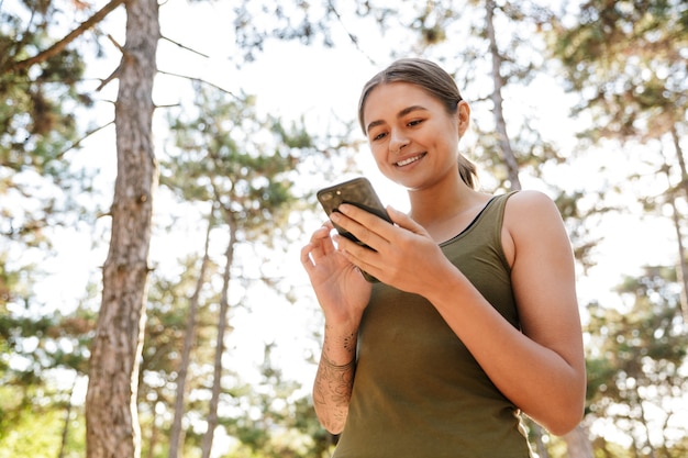  happy caucasian woman in sportswear using cellphone and smiling while working out in sunny green park