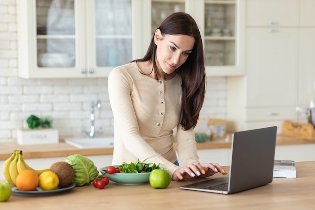 Felice donna caucasica in piedi nella cucina di casa con laptop e set di cibo sano, cerca su internet ricette per cucinare cibi dietetici