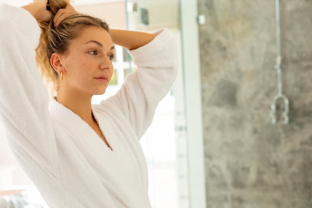 Photo happy caucasian woman wearing robe, tying up hair in bathroom. spending quality time alone at home.