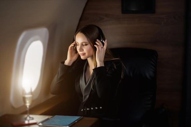 세련된 검은 드레스를 입은 행복한 백인 젊은 금발 여성은 개인 제트기에서 비행 중 휴식을 취하고 눈을 감고 무선 헤드폰을 사용하여 음악을 듣습니다.