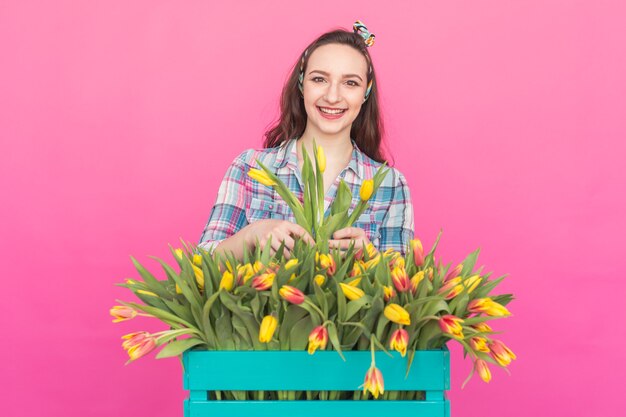 Happy caucasian young woman with box of yellow tulips on pink surface