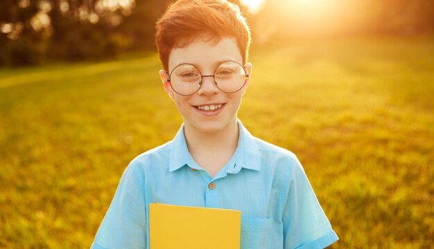 Happy charismatic schoolboy with ginger hair in blue shirt and round eyeglasses, smiling and looking at camera while standing on grassy meadow with notebook in hand on sunny day