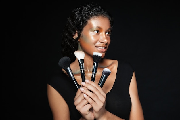 Happy charming american young woman standing and holding makeup brushes