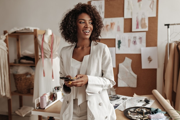 Happy charming dark-skinned curly brunette woman in white jacket, stylish top smiles sincerely, looks away, holds phone