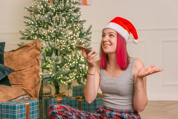 Happy charming young lady sits on the floor communicates on the phone against the background of a Christmas tree with gifts