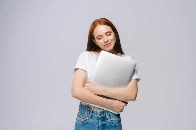 Happy charming young woman student holding laptop computer with closed eyes on isolated gray background. Pretty lady model with red hair emotionally showing facial expressions in studio, copy space.