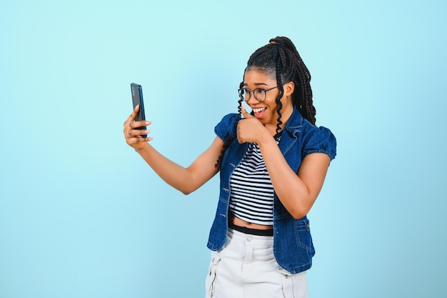 Happy cheerful afro american woman looks at screen of smart\
phone enjoys online chatting types text message surfs social\
networks dressed casually poses against blue background technology\
concept