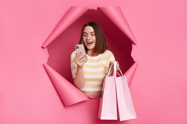 Happy cheerful amazed brunette woman wearing striped t shirt posing in torn pink paper wall holding shopping bags using smartphone ordering purchases online