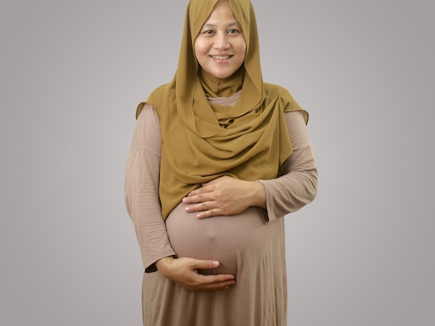 Happy cheerful Asian muslim pregnant mother wearing hijab smiling while holding her big belly