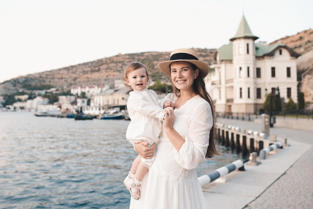 Happy cheerful baby girl sitting on mother hands wear stylish white dresse and straw hat relax over sea shore at quayside outdoors close up. Looking at camera. Motherhood. Maternity.