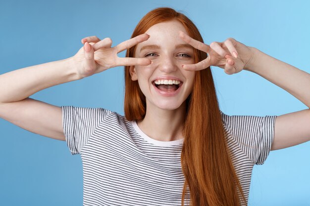 Foto felice allegro spensierato caucasica ragazza rossa zenzero capelli lunghi divertirsi esprimere felicità gioia amichevole atteggiamento mostra pace vittoria gesto discoteca segni occhi ridendo, in piedi sfondo blu.