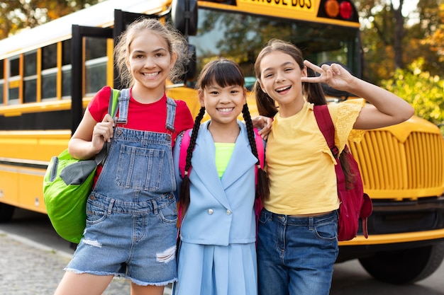 Счастливые веселые одноклассницы, позирующие вместе на улице возле желтого школьного автобуса