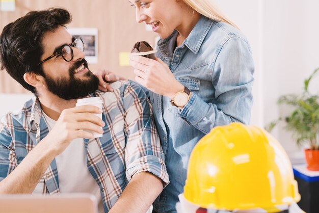 Happy cheerful couple of engineer coworkers having a break with a muffin and coffee in a paper cup in the office.