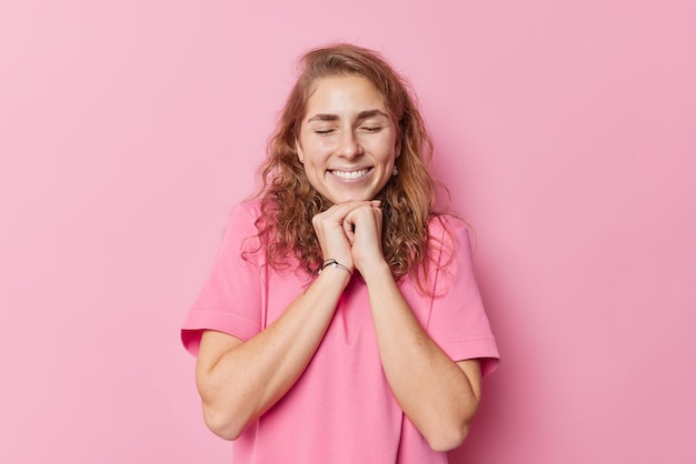 Happy cheerful European woman with dreamy expression has long wavy hair perfect teeth keeps hands under chin dressed in casual t shirt isolated over pink background. Positive emotions concept