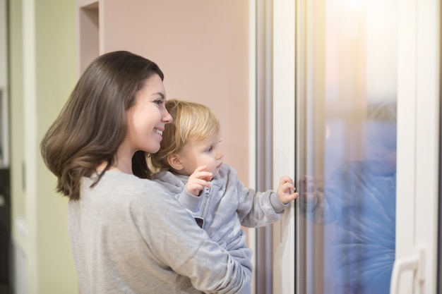 Happy cheerful family lifestyle Young smiling Mother hugging her baby and looking at window