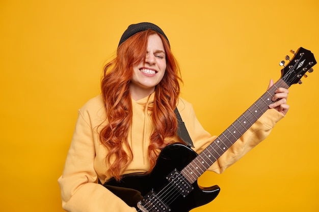 Happy cheerful female teenager has natual long red hair plays black electric guitar in professional studio wears hat casual hoodie 