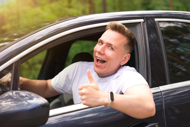 Felice ragazzo allegro conducente giovane uomo che guida la sua auto sorridente mostra pollice in su dal finestrino dell'automobile