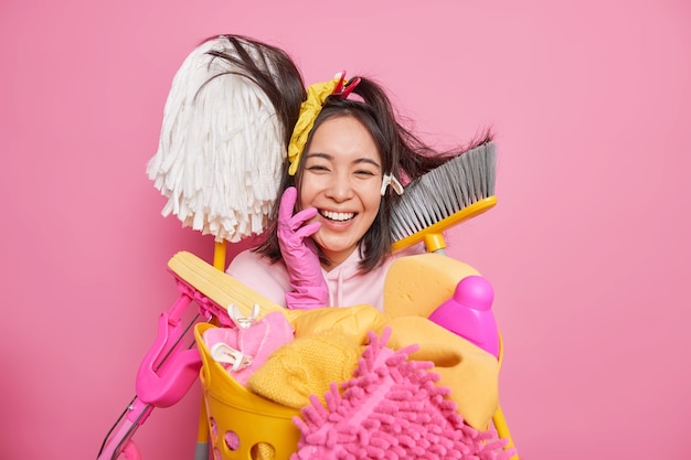 Happy cheerful housewife foolishes around while doing house cleanup surrounded by cleaning supplies basket full of dirty clothes to wash isolated over pink background. Household laundering concept