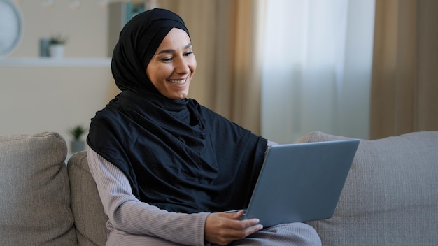 Happy cheerful islamic young girl making video call sit in living room enjoy pleasant online