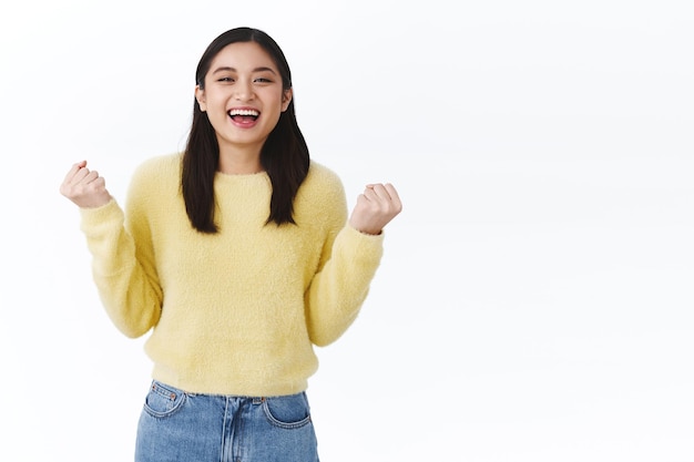 Felice allegra fortunata ragazza asiatica che vince, diventa campione e gioisce, stringe i pugni e li gonfia di gioia, dicendo evviva sì e sorridendo