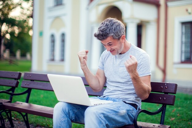 Uomo allegro felice con un computer portatile che si siede nel parco. concetto di persone, tecnologia e stile di vita