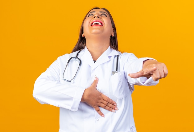 Happy and cheerful middle age woman doctor in white coat with stethoscope laughing out pointing with index finger at something