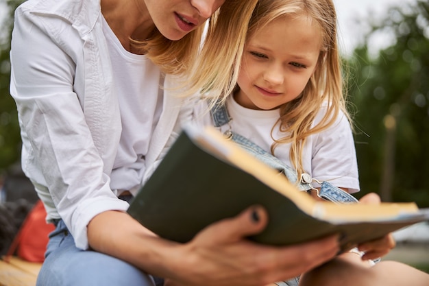 Happy cheerful miss resting with mother in the park while reading new fairytale of princess