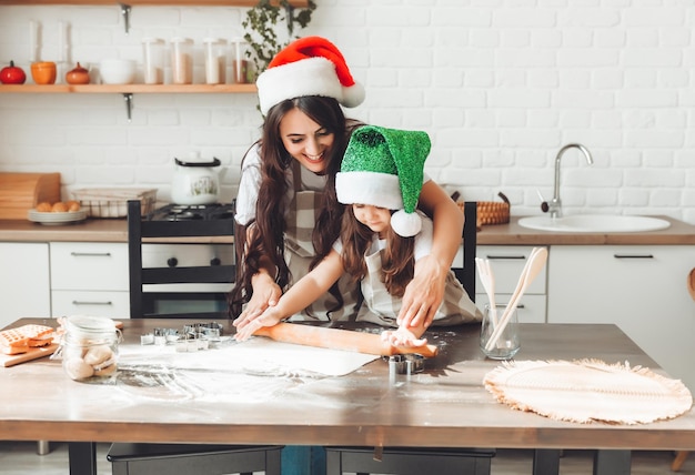 サンタ クロースの帽子をかぶった幸せな陽気な母と子は、新年とクリスマスのキッチンでクリスマス クッキーを作っています。