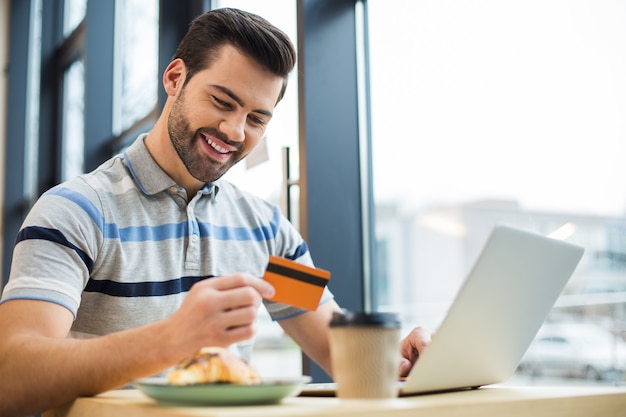 Счастливый веселый приятный человек улыбается и смотрит на свою кредитную карту, сидя перед ноутбуком