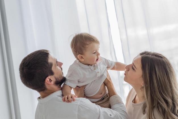 Happy cheerful parents having fun with cute child son cuddling playing near window