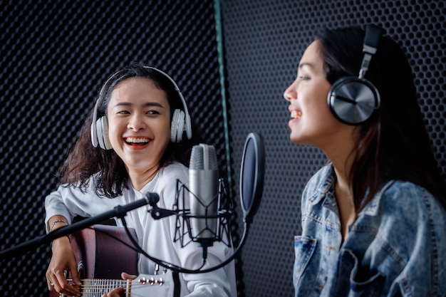 Happy cheerful pretty smiling of portrait Two young Asian woman vocalist Wearing Headphones with a guitar recording a song front of microphone in a professional studio