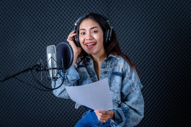 Happy cheerful pretty smiling of portrait of young Asian woman vocalist Wearing Headphones recording a song front of microphone in a professional studio