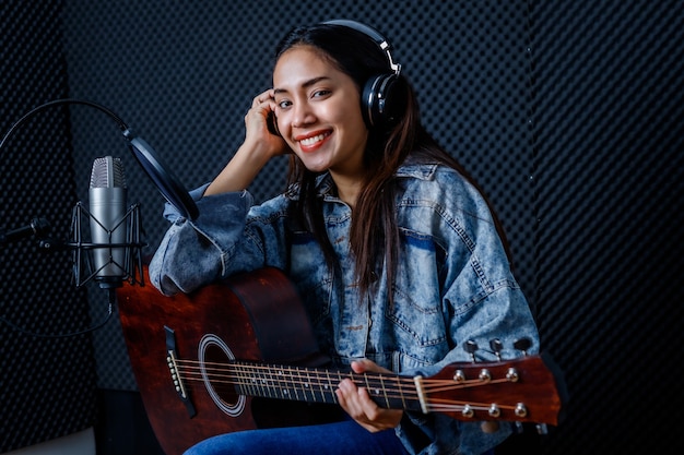 Felice allegro piuttosto sorridente del ritratto di una giovane cantante asiatica che indossa le cuffie con una chitarra che registra una canzone davanti al microfono in uno studio professionale