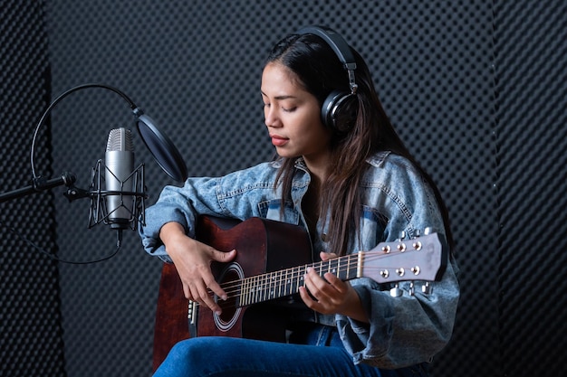 Photo happy cheerful pretty smiling of portrait a young asian woman vocalist wearing headphones with a guitar recording a song front of microphone in a professional studio