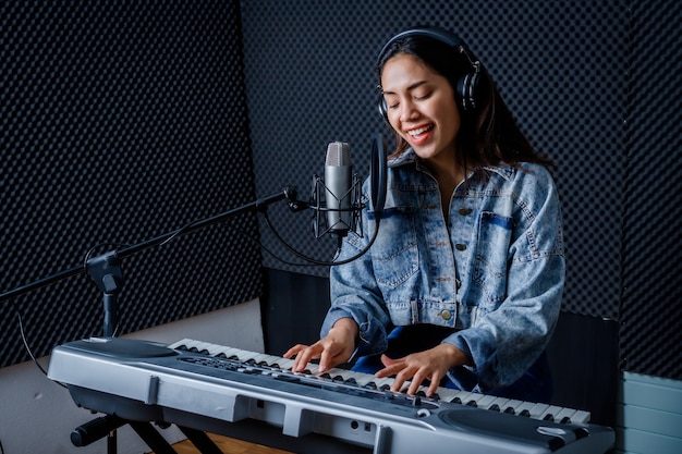 Happy cheerful pretty smiling of young Asian woman vocalist Wearing Headphones recording a song front of microphone and playing the keyboard during rehearsal of her band in a professional studio