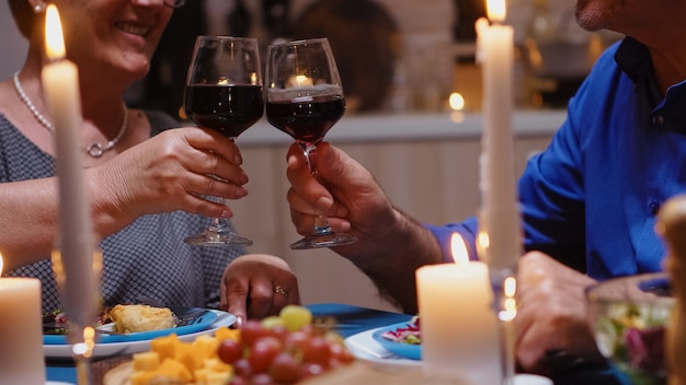 Happy cheerful senior elderly couple dining together with red wine in the cozy kitchen. Aged retired old people enjoying the meal, celebrating their anniversary in the dining room.