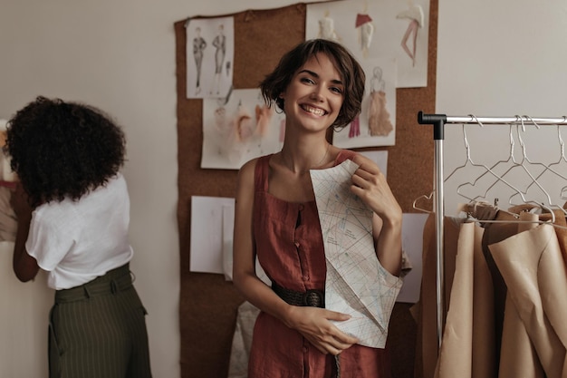 Happy cheerful shorthaired woman in linen red dress smiles sincerely and holds clothes pattern Young curly woman designs dress