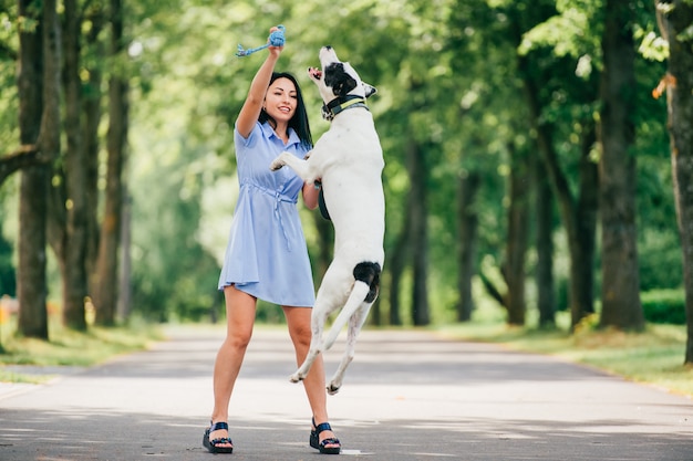 Happy cheerful smiling brunette girl in blue summer dress playing with big hunting dog in park