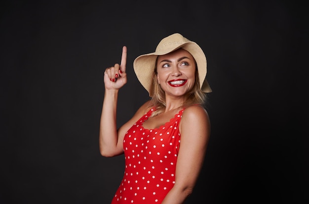 Happy cheerful stunning blonde Caucasian woman in red outfit with white polka dots and straw summer hat smiles beautiful white toothy smile pointing up finger at copy space on black background