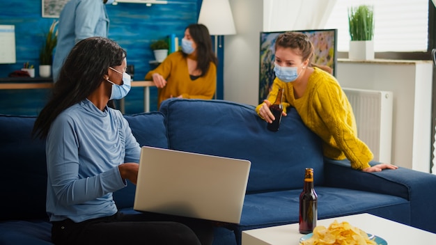 Happy cheerful woman holding beer bottle looking at laptop talking with african friend in living room wearing face mask to prevent coronavirus spread in time of global pandemic. Conceptual footage.