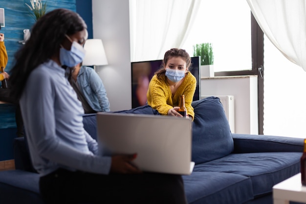 Happy cheerful woman holding beer bottle looking at laptop talking with african friend in living room wearing face mask to prevent coronavirus spread in time of global pandemic. Conceptual footage.