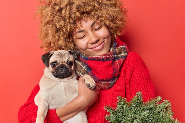 Happy cheerful woman tilts head smiles gently keeps eyes closed embraces pug dog with love carries green spruce wreath wears sweater and winter scarf around neck isolated over red background