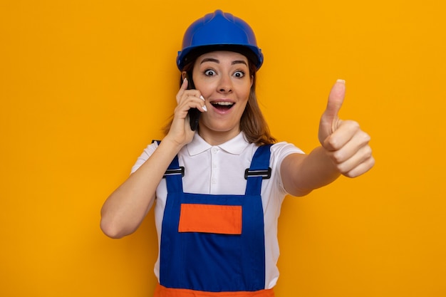 Happy and cheerful young builder woman in construction uniform and safety helmet looking while talking on mobile phone showing thumbs up